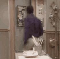 a man in a purple shirt is standing in a hallway next to a table with utensils on it .