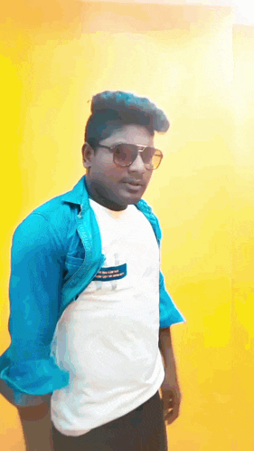 a man wearing sunglasses and a blue shirt is standing in front of a yellow wall