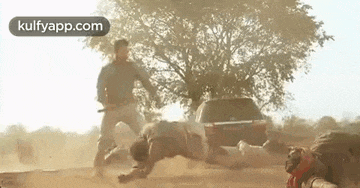 a group of men are fighting each other on a dirt road in front of a car .