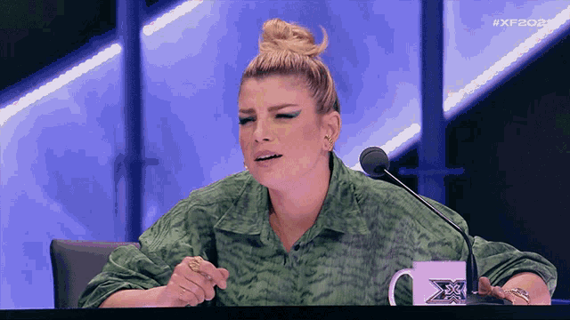 a woman in a green shirt is sitting at a table with a microphone and a mug that says x factor