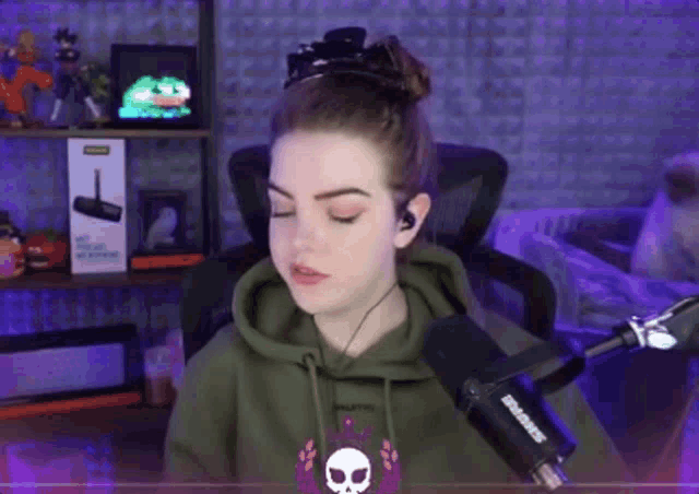 a woman wearing a green hoodie with a skull on it stands in front of a shure microphone .
