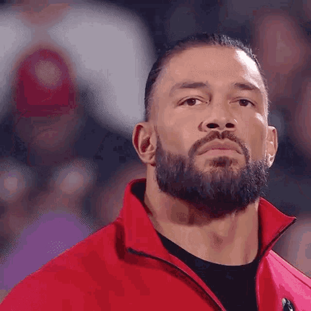 roman reigns is wearing a red jacket and a black shirt and looking at the camera .