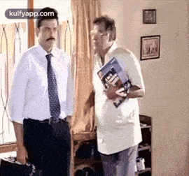 two men in suits and ties are standing next to each other in a living room . one man is holding a book .