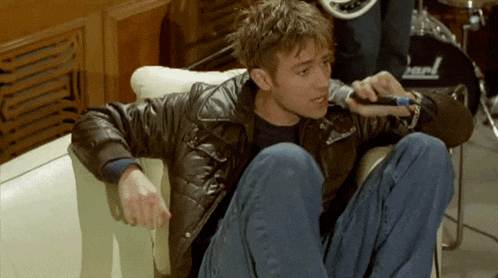 a man in a leather jacket sits on a couch holding a microphone in front of a drum set