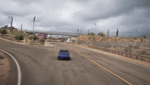 a blue car is driving down a desert road with a bridge in the background