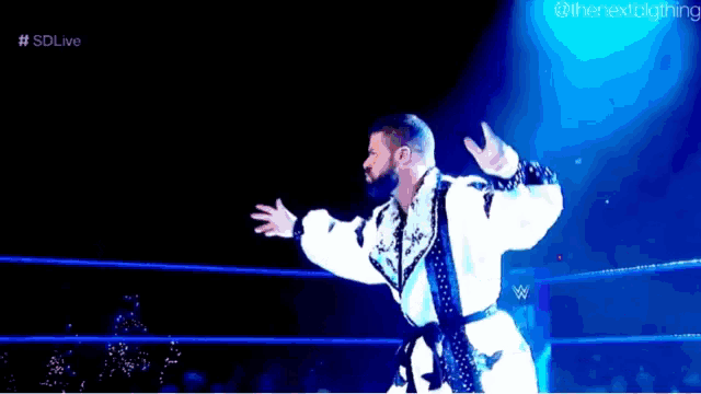 a wrestler in a white robe is standing in a wrestling ring with his arms outstretched .