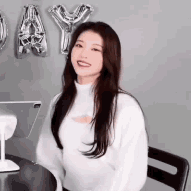 a woman in a white sweater is sitting at a table with balloons behind her that say happy day