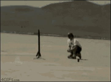 a man is kneeling on the ground in the desert while another man takes a picture of a plane flying overhead .