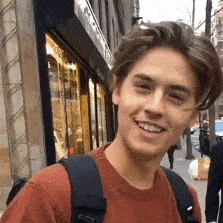 a young man wearing a red sweater and a backpack is smiling on the street .