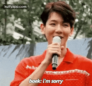a young man in a red shirt is holding a microphone and saying `` baek , i 'm sorry ''