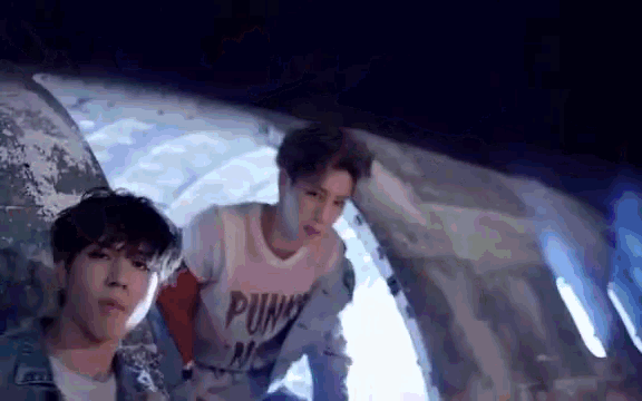 two young men are standing next to each other in front of an airplane . one of the men is wearing a punk shirt .