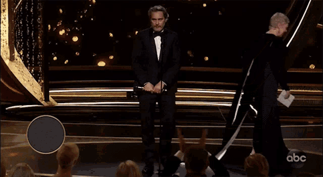 a man in a tuxedo stands on a stage in front of a sign that says " best actor 2020 "