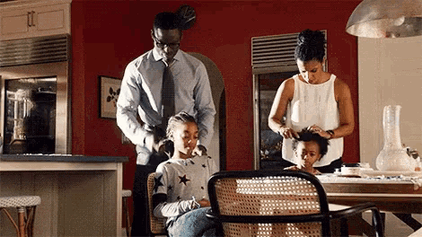 a family is sitting at a table in a kitchen while a man and a woman are combing their hair .