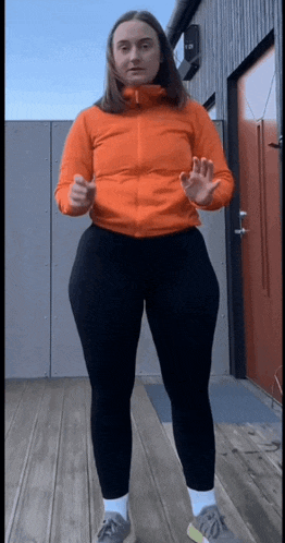 a woman in an orange jacket and black leggings stands on a wooden deck