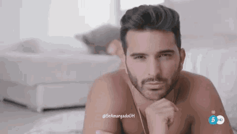 a shirtless man with a beard is laying on a bed with his hand on his chin .