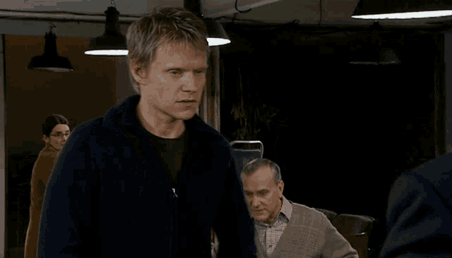 a man in a blue sweater stands in a dark room with other men
