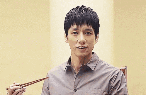 a man in a grey shirt is sitting in a chair holding chopsticks .