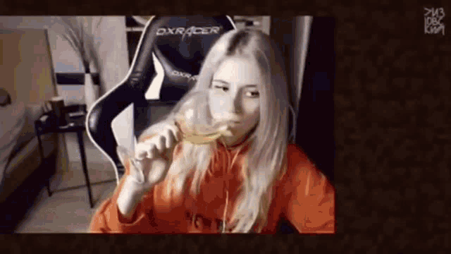 a woman in an orange hoodie is sitting in a dxracer chair eating a piece of cheese .
