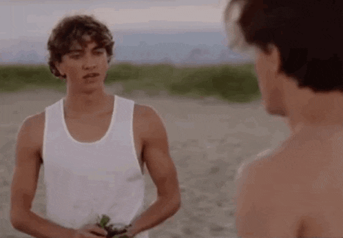 a man in a white tank top is standing next to another man without a shirt on the beach .