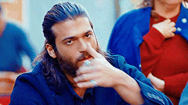 a man with long hair and a beard is holding his finger to his nose while sitting at a table .