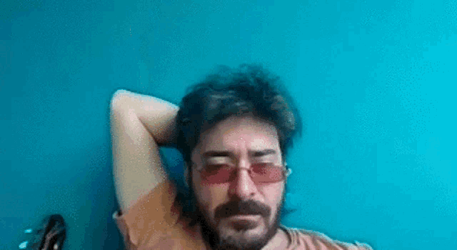 a man with a beard and red sunglasses is sitting in front of a blue wall with his hands behind his head .