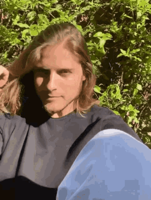 a man with long hair and a beard is standing in front of a bush