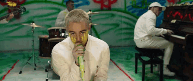 a man with yellow paint on his face is singing into a microphone while another man plays the piano