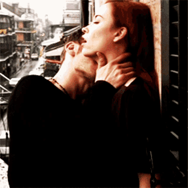 a man and a woman are kissing on a balcony with a city in the background