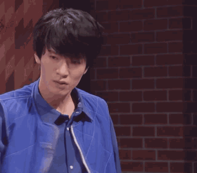 a young man wearing a blue vest and a blue shirt stands in front of a brick wall