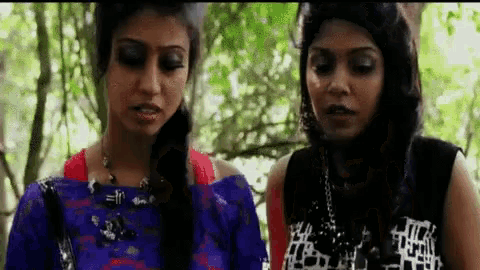 two women are standing next to each other in a forest and talking .