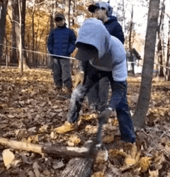 a man wearing a hooded jacket is cutting a log in the woods .