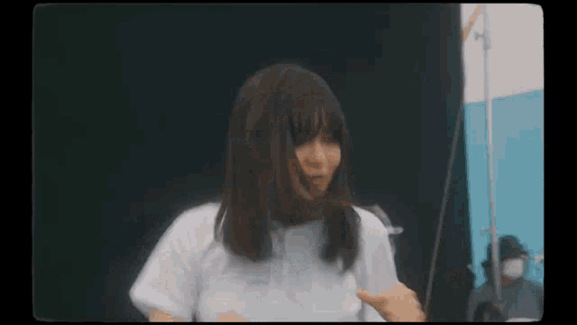 a woman in a white shirt is standing in front of a black wall and looking at the camera .