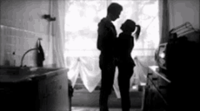 a black and white photo of a man and a woman hugging each other in front of a window .