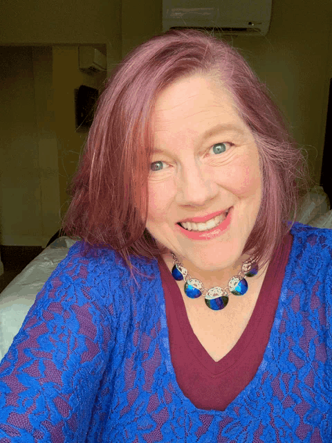 a woman with pink hair is smiling and wearing a blue top and necklace