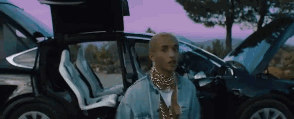 a man in a denim shirt and gold chains is standing in front of a car with the trunk open .