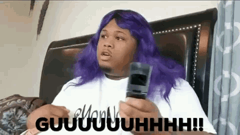 a man with purple hair is sitting on a bed holding a cell phone and saying guuuuhhhh !!