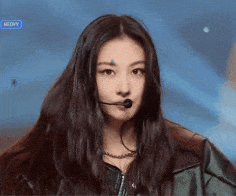a woman with long hair is wearing a microphone and the word meovv is on the screen behind her