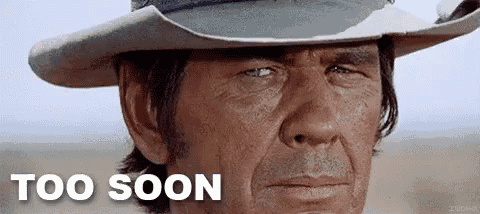 a man wearing a cowboy hat is looking at the camera with the words `` too soon '' written on the bottom .