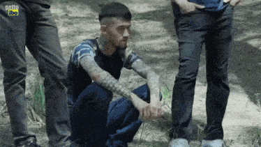 a man with a tattoo on his arm is squatting down in the dirt while two other men stand behind him .