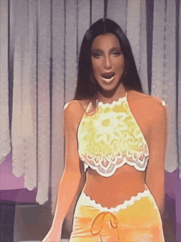 a woman in a yellow crop top and orange shorts is standing in front of a curtain .