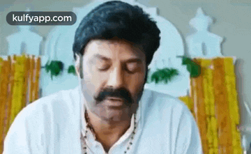 a man with a mustache and a white shirt is sitting in front of a wall with his eyes closed .