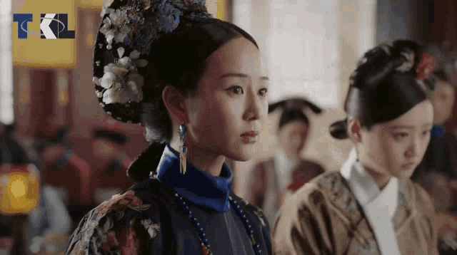 a woman in a blue and gold dress with a flower in her hair stands next to another woman