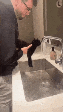 a man is petting a black cat that is standing in a sink