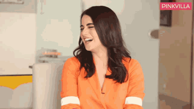 a woman in an orange jacket is laughing with a pinkvilla logo in the background