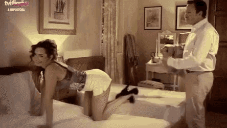 a woman is kneeling on a bed while a man holds a stuffed animal in front of a tv screen