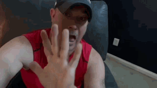 a man wearing a hat and a red tank top is making a stop sign with his hand .