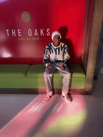 a man sits on a green bench in front of a red wall that says the oaks collective