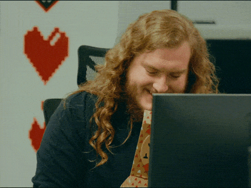a man with long red hair is smiling while looking at his laptop
