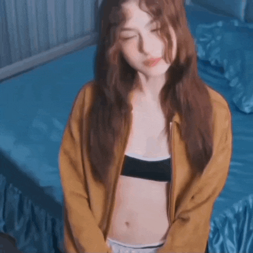 a woman in a bra and a jacket is sitting on a bed with her eyes closed .