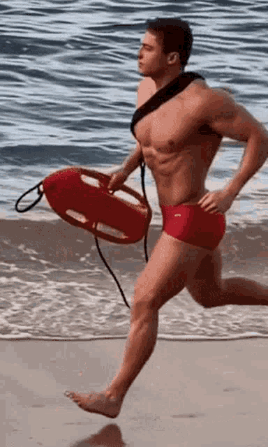 a shirtless lifeguard is running on the beach holding a life preserver .
